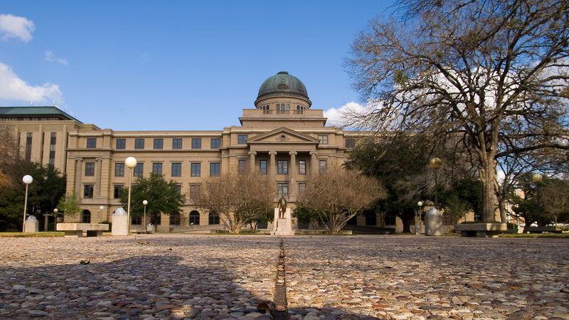 The domed Academic Building at Texas A&M University in College Station, seen across the cobbled plaza with bare oak trees under a blue sky.