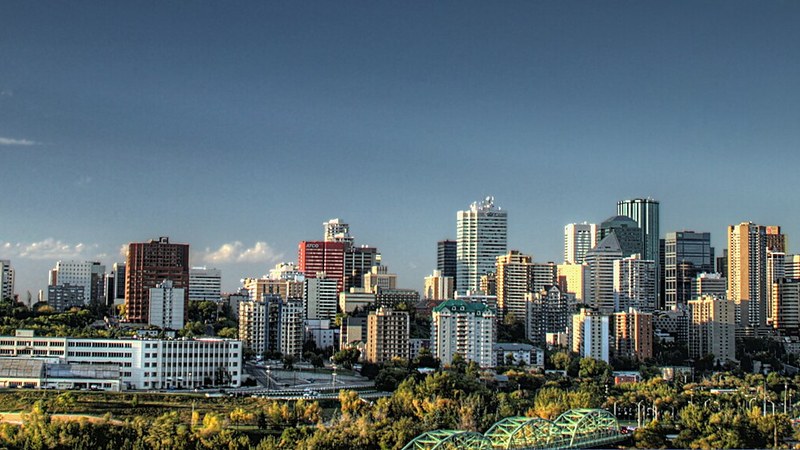 Edmonton skyline above the river valley, with autumn trees and a green steel bridge in the foreground.