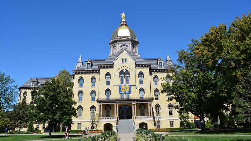 The Main Building of the University of Notre Dame with its Golden Dome topped by a statue of Mary, framed by trees on a sunny day, at Notre Dame next to South Bend, Indiana.