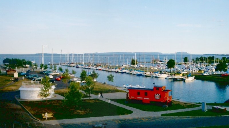 Sailboats in Thunder Bay marina beside waterfront paths and a red railway caboose.