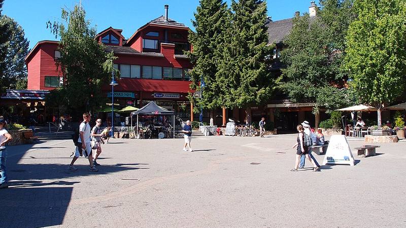Sunny pedestrian plaza in Whistler Village, with chalet-style buildings, trees and outdoor cafes.